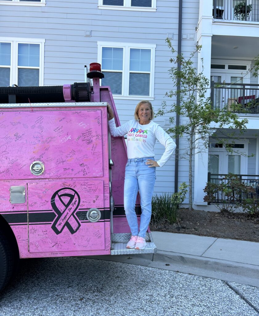 A woman wearing a shirt that says "fight cancer in all colors" stands on the back of a pink truck with a cancer ribbon on it