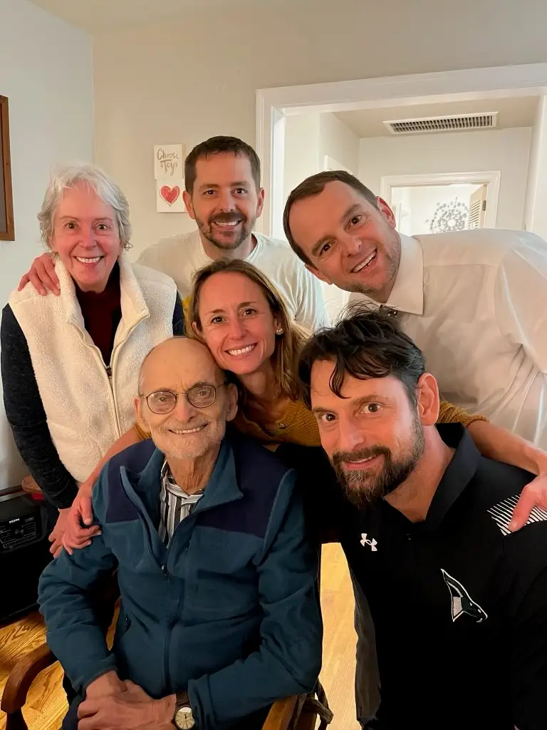 A group of six people gathered closely indoors, smiling for a photo. An older person wearing glasses and a blue jacket sits in front, with five others standing behind them,