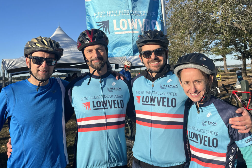 Four people pose for a photo in cycling gear with a flag behind that says Lowvelo.