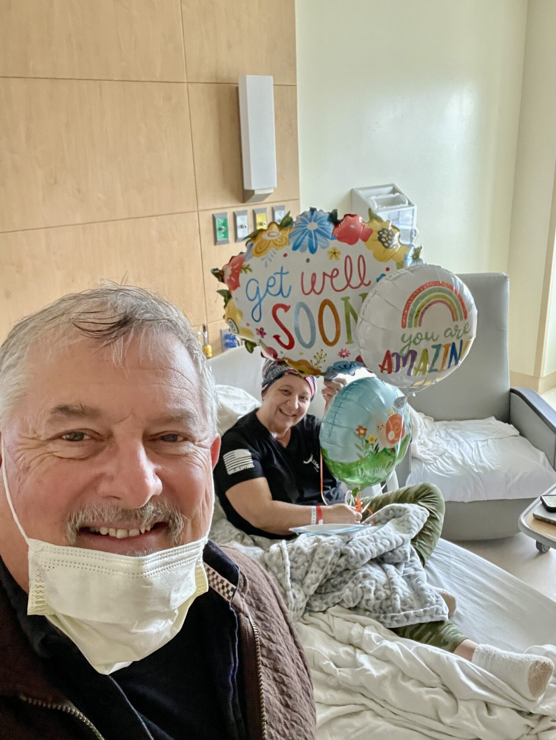 Two people take a selfie. One is in a hospital bed with get well balloons.