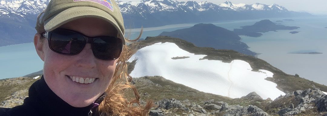 Lucy Boyce hikes in Alaska with water and mountains behind her
